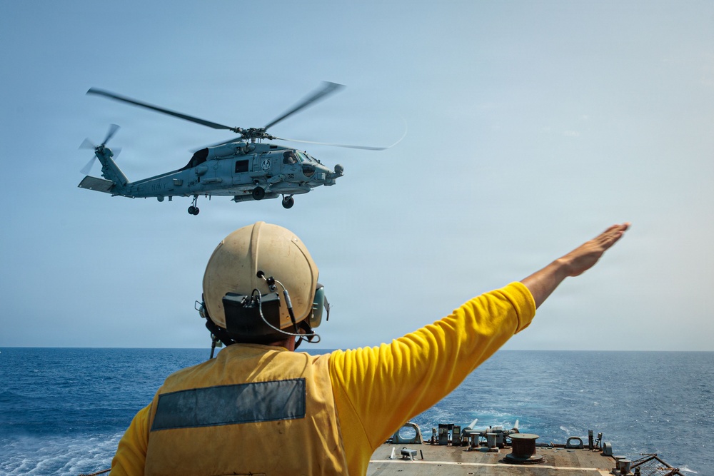 A naval deck crew member in a yellow jersey and flight deck helmet is signaling with an outstretched arm toward a military helicopter hovering above a warship's deck at sea. The helicopter appears to be a Sikorsky MH-60R Seahawk, commonly used by the U.S. Navy for maritime operations. The scene depicts a standard flight deck operations procedure, likely guiding the helicopter for landing or departure.