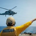 A naval deck crew member in a yellow jersey and flight deck helmet is signaling with an outstretched arm toward a military helicopter hovering above a warship's deck at sea. The helicopter appears to be a Sikorsky MH-60R Seahawk, commonly used by the U.S. Navy for maritime operations. The scene depicts a standard flight deck operations procedure, likely guiding the helicopter for landing or departure.