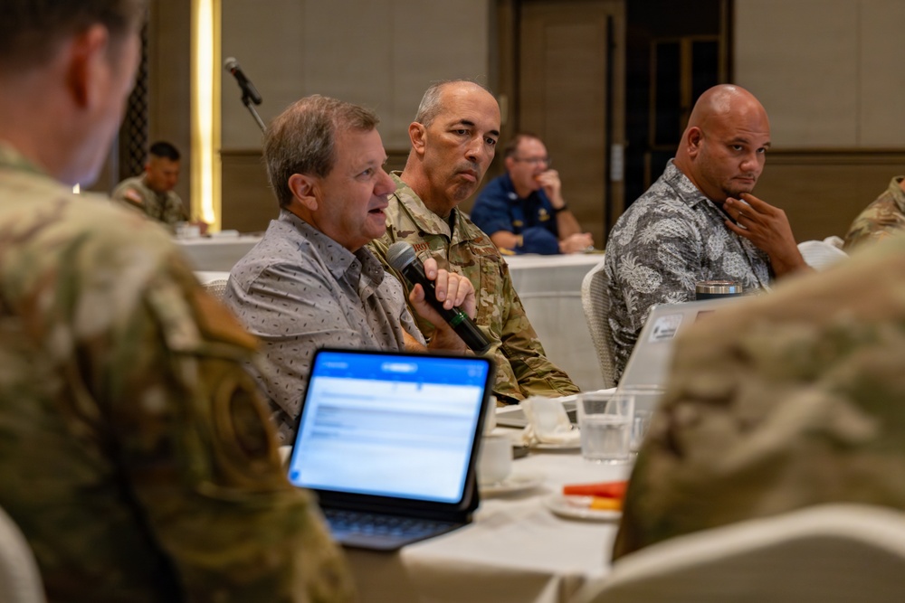 A formal meeting or conference is taking place in what appears to be a hotel ballroom or conference facility. Multiple individuals, including military personnel in camouflage uniforms and civilians, are seated at round tables. A civilian man is speaking into a microphone while a soldier in uniform listens attentively beside him.