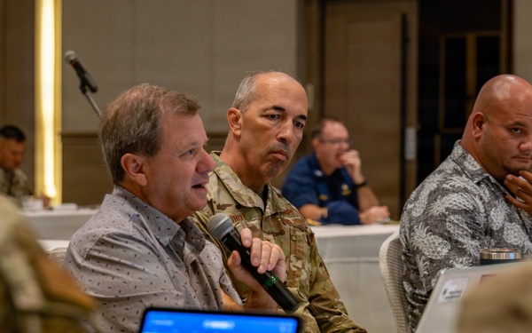 A formal meeting or conference is taking place in what appears to be a hotel ballroom or conference facility. Multiple individuals, including military personnel in camouflage uniforms and civilians, are seated at round tables. A civilian man is speaking into a microphone while a soldier in uniform listens attentively beside him.