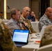 A formal meeting or conference is taking place in what appears to be a hotel ballroom or conference facility. Multiple individuals, including military personnel in camouflage uniforms and civilians, are seated at round tables. A civilian man is speaking into a microphone while a soldier in uniform listens attentively beside him.