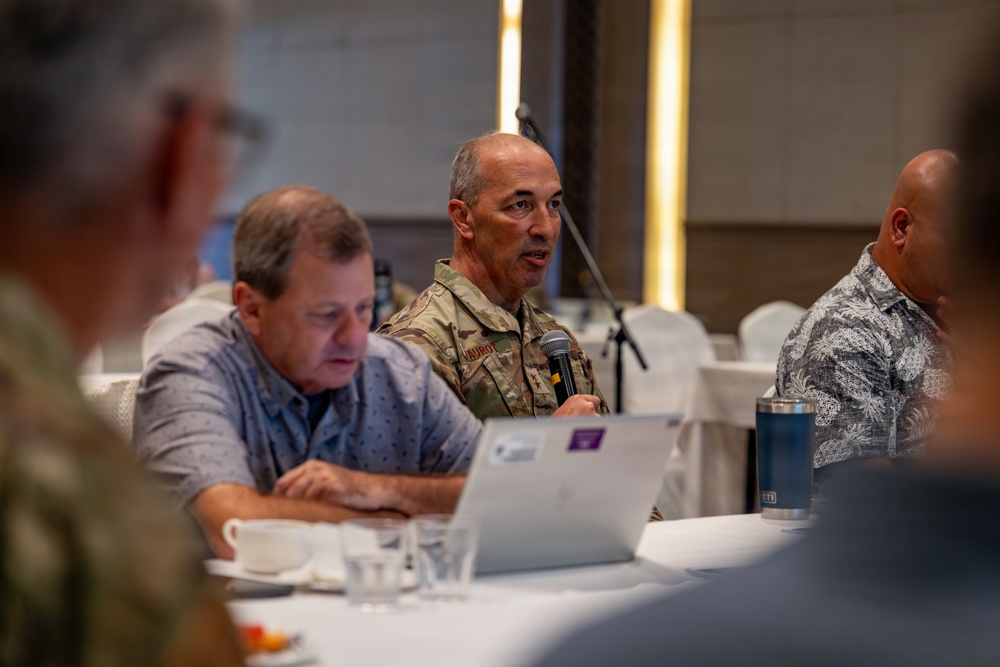 A military officer in camouflage uniform is speaking into a microphone at a conference or meeting table, flanked by civilians in casual attire. The setting appears to be a formal meeting room or hotel conference space with white tablecloths. The officer's name tape reads 'VAUROT' and rank insignia suggesting a general officer grade is partially visible.