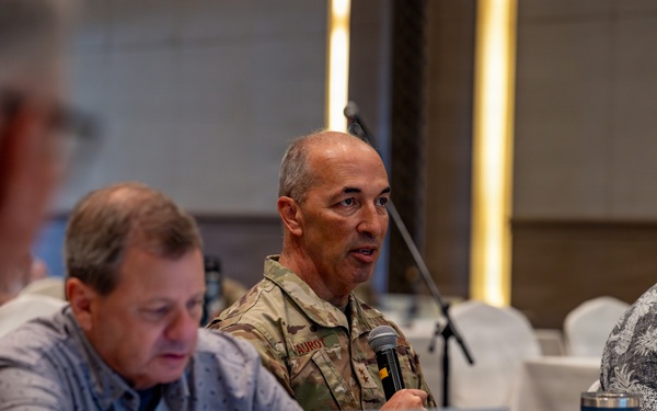 A military officer in camouflage uniform is speaking into a microphone at a conference or meeting table, flanked by civilians in casual attire. The setting appears to be a formal meeting room or hotel conference space with white tablecloths. The officer's name tape reads 'VAUROT' and rank insignia suggesting a general officer grade is partially visible.