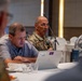 A military officer in camouflage uniform is speaking into a microphone at a conference or meeting table, flanked by civilians in casual attire. The setting appears to be a formal meeting room or hotel conference space with white tablecloths. The officer's name tape reads 'VAUROT' and rank insignia suggesting a general officer grade is partially visible.