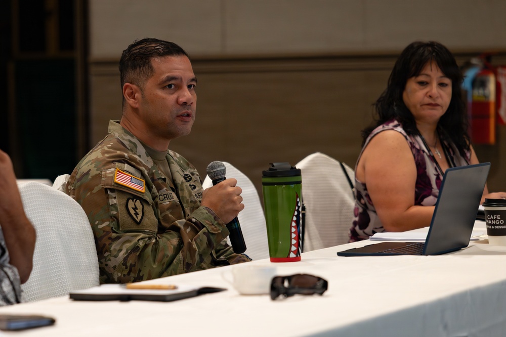 A U.S. Army soldier in OCP (Operational Camouflage Pattern) uniform is speaking into a microphone at a conference or meeting table. A civilian woman seated beside him appears to be listening while a laptop is open in front of her. The setting appears to be a formal meeting or briefing in a hotel or conference facility.