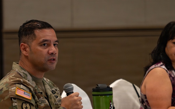 A U.S. Army soldier in OCP (Operational Camouflage Pattern) uniform is speaking into a microphone at a conference or meeting table. A civilian woman seated beside him appears to be listening while a laptop is open in front of her. The setting appears to be a formal meeting or briefing in a hotel or conference facility.