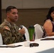 A U.S. Army soldier in OCP (Operational Camouflage Pattern) uniform is speaking into a microphone at a conference or meeting table. A civilian woman seated beside him appears to be listening while a laptop is open in front of her. The setting appears to be a formal meeting or briefing in a hotel or conference facility.