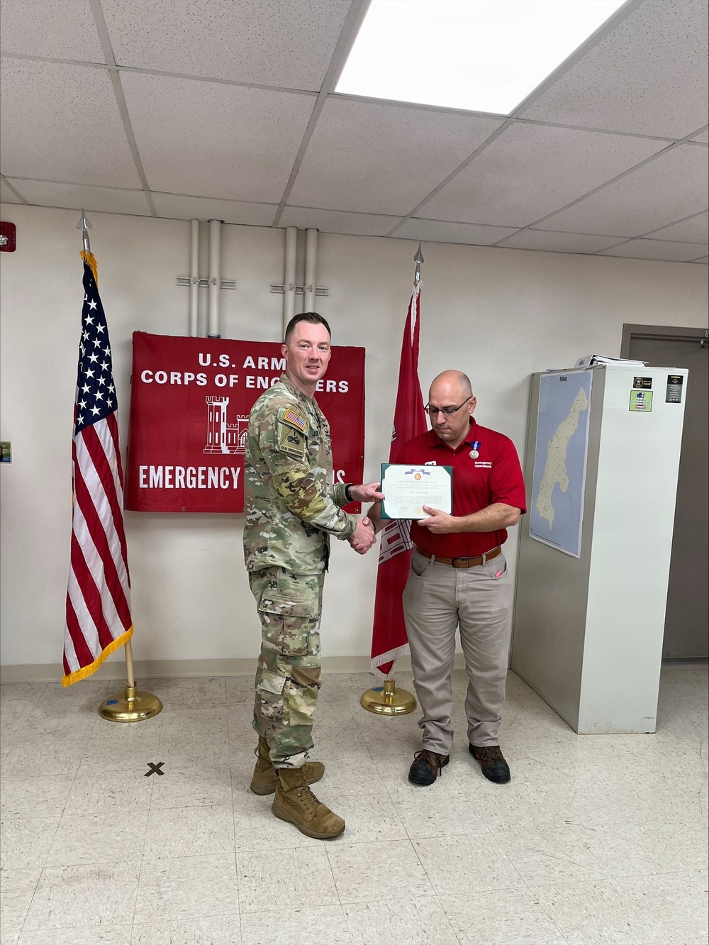A U.S. Army soldier in OCP (Operational Camouflage Pattern) uniform is presenting a certificate or award to a civilian employee wearing a red polo shirt with a medal pinned to it. The ceremony is taking place in front of a U.S. Army Corps of Engineers Emergency Operations banner, flanked by the U.S. flag and the USACE castle flag. This appears to be a formal recognition or award ceremony.
