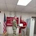 A U.S. Army soldier in OCP (Operational Camouflage Pattern) uniform is presenting a certificate or award to a civilian employee wearing a red polo shirt with a medal pinned to it. The ceremony is taking place in front of a U.S. Army Corps of Engineers Emergency Operations banner, flanked by the U.S. flag and the USACE castle flag. This appears to be a formal recognition or award ceremony.