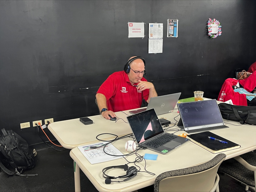 A man wearing a red polo shirt and headphones sits at a table with multiple laptops and devices, appearing to be working in a sports operations or broadcasting setup. A baseball is visible on the table among papers, cables, and electronic equipment. The setting appears to be a back-room or press area at a baseball venue, indicated by the dark walls and posted documents.