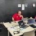 A man wearing a red polo shirt and headphones sits at a table with multiple laptops and devices, appearing to be working in a sports operations or broadcasting setup. A baseball is visible on the table among papers, cables, and electronic equipment. The setting appears to be a back-room or press area at a baseball venue, indicated by the dark walls and posted documents.