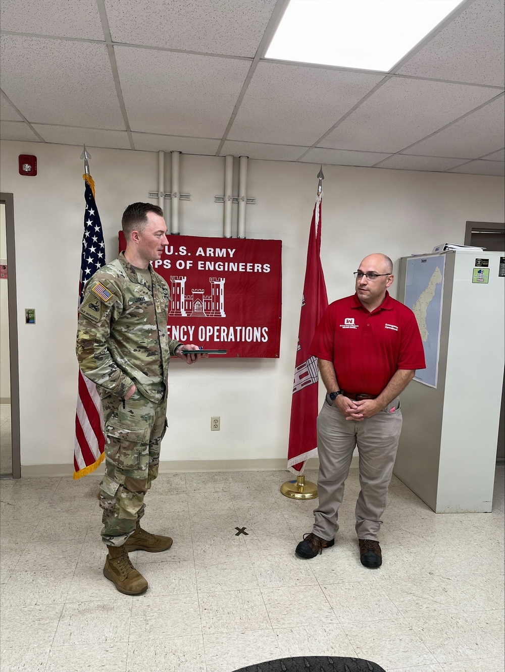 Two individuals stand in an official U.S. Army Corps of Engineers Emergency Operations room. One person is wearing U.S. Army combat uniform (OCP/multicam) and the other is wearing a red polo shirt with what appears to be a U.S. Army Corps of Engineers logo. They appear to be engaged in a formal or semi-formal interaction, possibly a briefing or recognition ceremony.