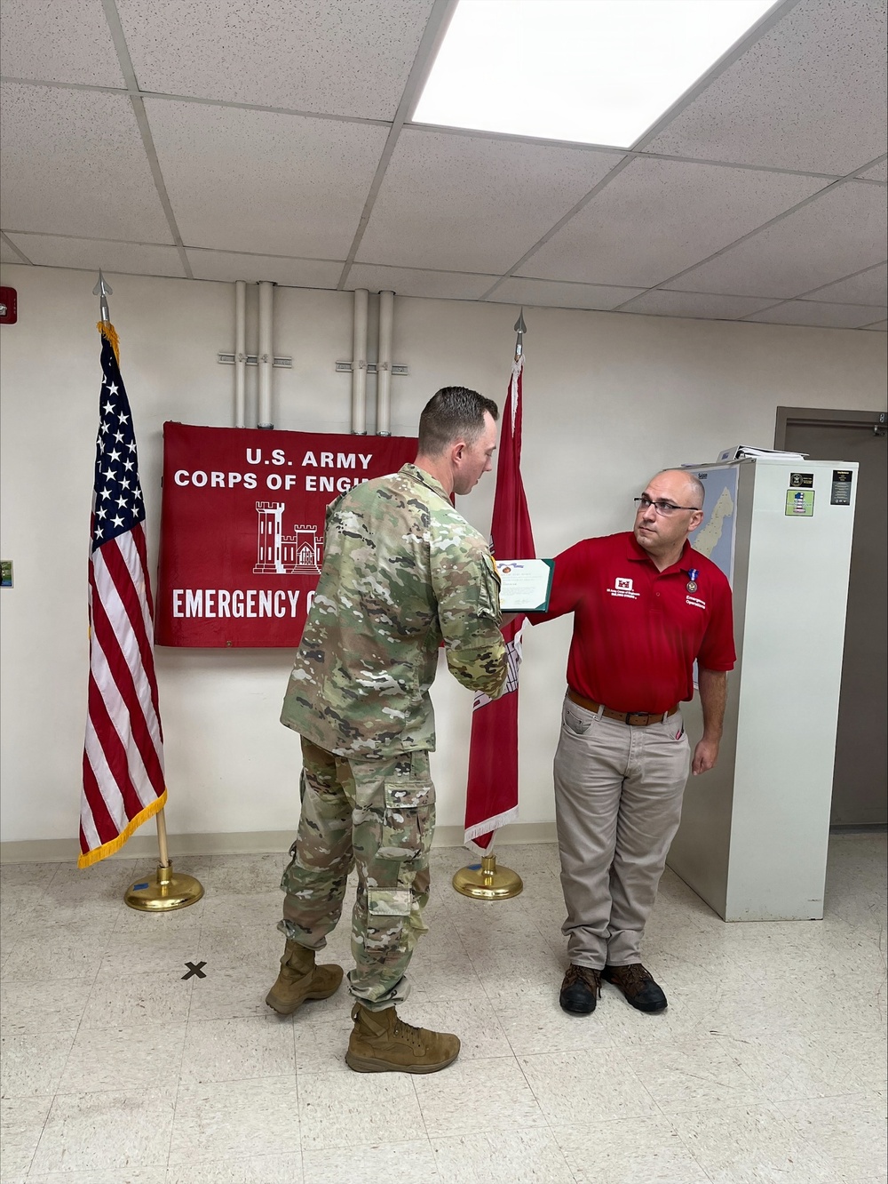 A U.S. Army soldier in OCP camouflage uniform is presenting a certificate or document to a civilian man wearing a red polo shirt, in what appears to be an official recognition or award ceremony. The setting is an indoor office or operations room associated with the U.S. Army Corps of Engineers Emergency Operations. Two flags — the American flag and a red Army Corps of Engineers flag — flank the participants in front of a banner on the wall.