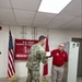 A U.S. Army soldier in OCP camouflage uniform is presenting a certificate or document to a civilian man wearing a red polo shirt, in what appears to be an official recognition or award ceremony. The setting is an indoor office or operations room associated with the U.S. Army Corps of Engineers Emergency Operations. Two flags — the American flag and a red Army Corps of Engineers flag — flank the participants in front of a banner on the wall.