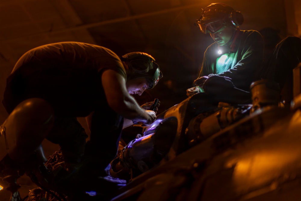 Two individuals are working in low-light conditions, using headlamps and handheld lights to perform what appears to be maintenance or repair work on mechanical equipment. The scene is illuminated primarily by artificial light sources including a bright headlamp worn by one person and a UV/blue-tinted light being used by the other. The orange ambient lighting and protective eyewear suggest an industrial or military maintenance environment.