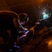 Two individuals are working in low-light conditions, using headlamps and handheld lights to perform what appears to be maintenance or repair work on mechanical equipment. The scene is illuminated primarily by artificial light sources including a bright headlamp worn by one person and a UV/blue-tinted light being used by the other. The orange ambient lighting and protective eyewear suggest an industrial or military maintenance environment.