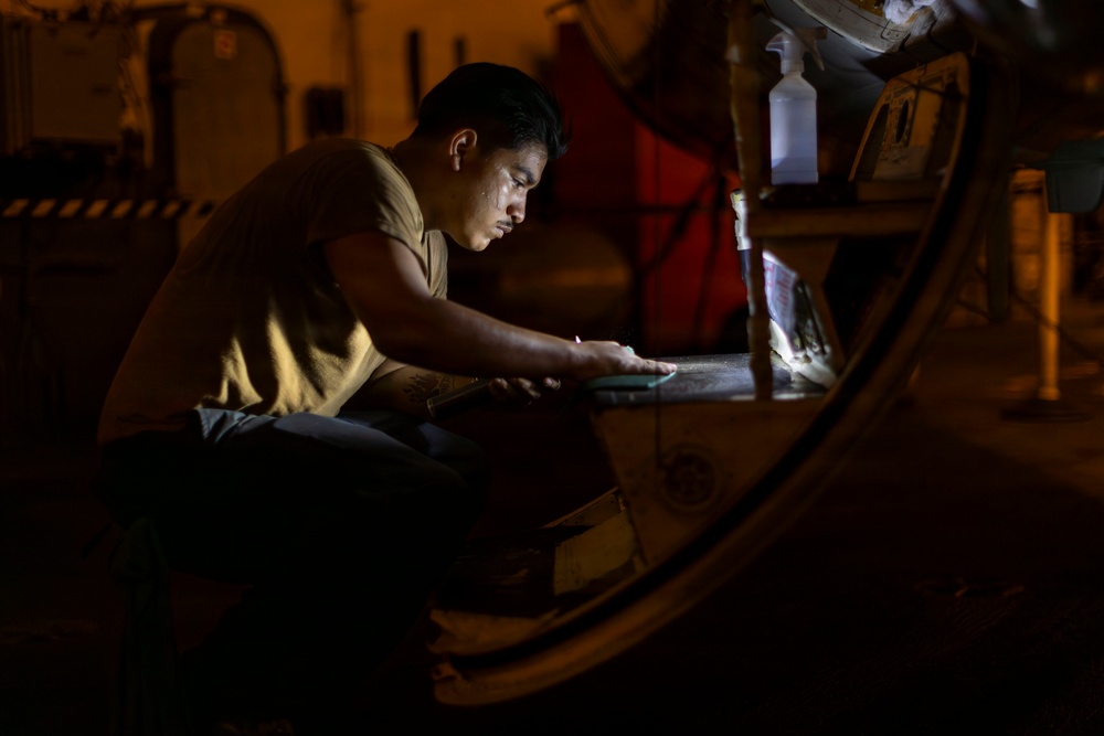 A young man in a tan/olive t-shirt is working intently in a dimly lit industrial or military environment, illuminated primarily by a small light source in front of him. He appears to be inspecting, cleaning, or performing maintenance on a large curved metal surface or component. The low-light conditions and focused posture suggest nighttime maintenance work.