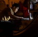 A young man in a tan/olive t-shirt is working intently in a dimly lit industrial or military environment, illuminated primarily by a small light source in front of him. He appears to be inspecting, cleaning, or performing maintenance on a large curved metal surface or component. The low-light conditions and focused posture suggest nighttime maintenance work.