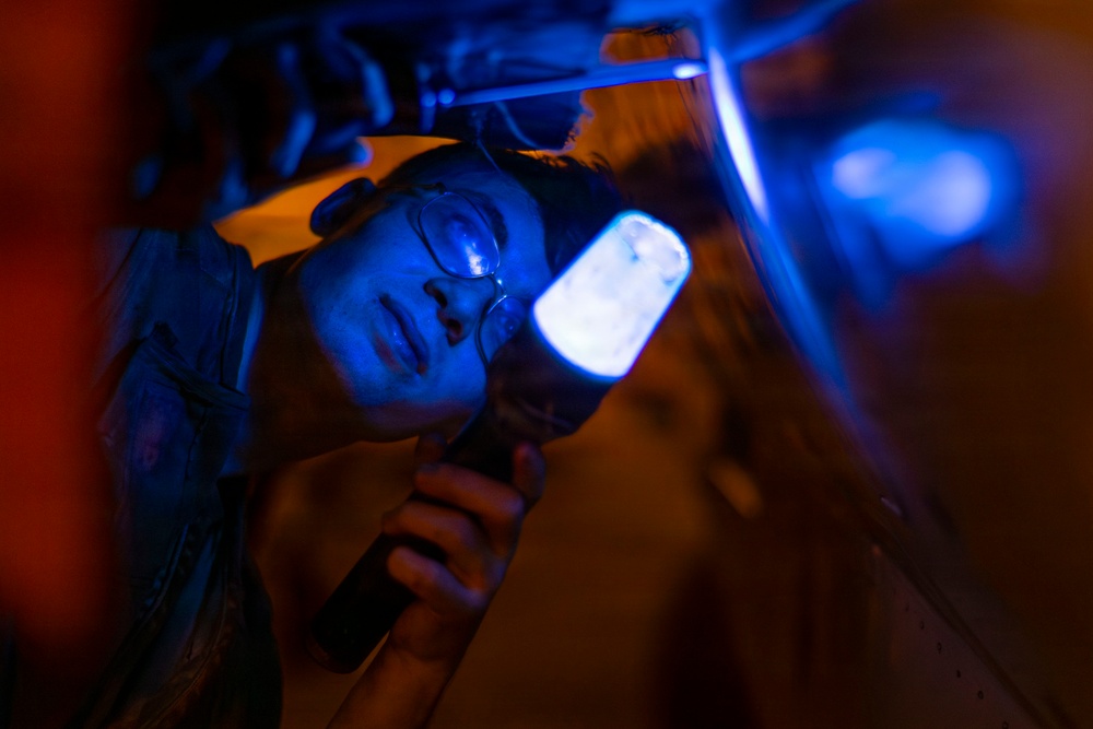 A person wearing glasses is lying on their back and holding a bright LED flashlight while inspecting the underside of a vehicle or machinery in a dark environment. The scene is dramatically lit with contrasting blue light from the flashlight and warm orange/red ambient light. The individual appears to be a mechanic or technician performing an inspection in low-light conditions.
