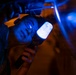 A person wearing glasses is lying on their back and holding a bright LED flashlight while inspecting the underside of a vehicle or machinery in a dark environment. The scene is dramatically lit with contrasting blue light from the flashlight and warm orange/red ambient light. The individual appears to be a mechanic or technician performing an inspection in low-light conditions.
