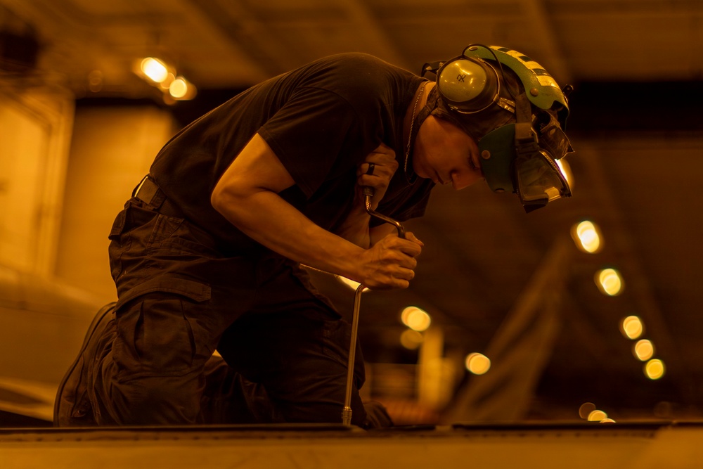 A technician or mechanic is working in what appears to be a large indoor facility, likely a hangar or industrial space, under warm amber lighting. The worker is kneeling and applying force to a tool, possibly a wrench or screwdriver, on a surface or piece of equipment. The individual is wearing appropriate personal protective equipment including hearing protection and a helmet with a face shield.