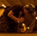 A technician or mechanic is working in what appears to be a large indoor facility, likely a hangar or industrial space, under warm amber lighting. The worker is kneeling and applying force to a tool, possibly a wrench or screwdriver, on a surface or piece of equipment. The individual is wearing appropriate personal protective equipment including hearing protection and a helmet with a face shield.