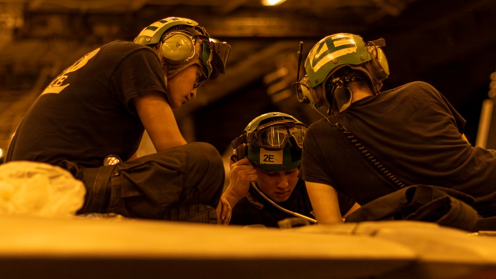 Three personnel wearing protective helmets with integrated hearing protection and goggles are gathered closely together, appearing to review documents or a surface in front of them. They are dressed in dark uniforms and their helmets are marked with identifiers including '2E'. The scene is illuminated by warm amber/orange lighting, suggesting a low-light operational environment such as a ship's hangar deck or flight deck.