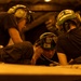 Three personnel wearing protective helmets with integrated hearing protection and goggles are gathered closely together, appearing to review documents or a surface in front of them. They are dressed in dark uniforms and their helmets are marked with identifiers including '2E'. The scene is illuminated by warm amber/orange lighting, suggesting a low-light operational environment such as a ship's hangar deck or flight deck.