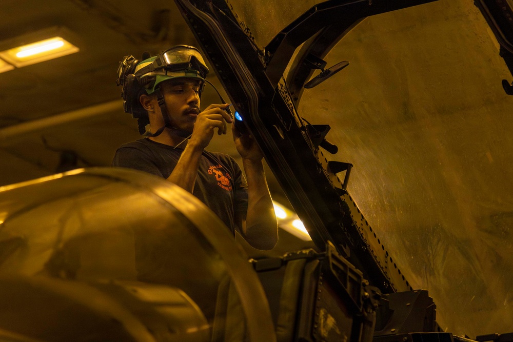 A technician or maintenance worker wearing a helmet with mounted equipment (likely night vision goggles or a headlamp) is performing close inspection or repair work on a large piece of equipment, possibly a military aircraft or vehicle. The work is being conducted in an enclosed, artificially lit space such as a hangar or maintenance bay. The worker appears to be using a small illuminated tool or device while examining a structural component.