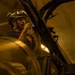 A technician or maintenance worker wearing a helmet with mounted equipment (likely night vision goggles or a headlamp) is performing close inspection or repair work on a large piece of equipment, possibly a military aircraft or vehicle. The work is being conducted in an enclosed, artificially lit space such as a hangar or maintenance bay. The worker appears to be using a small illuminated tool or device while examining a structural component.