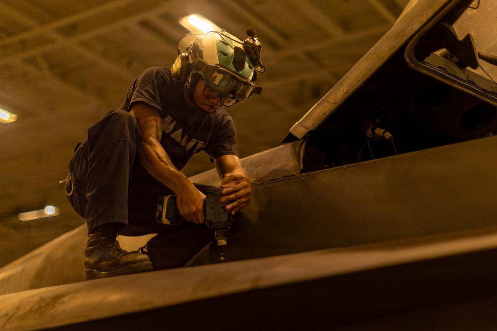 A military technician, likely a U.S. Navy sailor based on visible uniform text, is performing maintenance work on what appears to be a military aircraft in a hangar bay. The worker is crouched on top of the aircraft's surface, using a power tool near an open access panel. The image is taken from a low angle in warm artificial lighting, giving it a dramatic perspective.