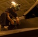 A military technician, likely a U.S. Navy sailor based on visible uniform text, is performing maintenance work on what appears to be a military aircraft in a hangar bay. The worker is crouched on top of the aircraft's surface, using a power tool near an open access panel. The image is taken from a low angle in warm artificial lighting, giving it a dramatic perspective.