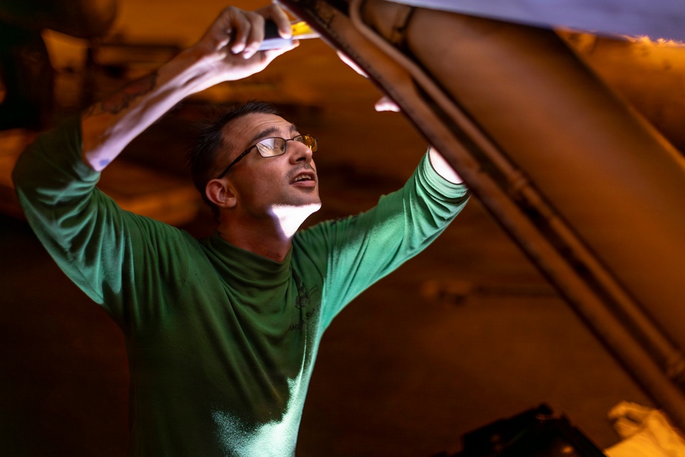 A man wearing glasses and a green long-sleeve shirt is working underneath or inside a large vehicle or aircraft, using a handheld light source to illuminate the area above him. He appears to be inspecting or performing maintenance on the underside of a large structure. The image is dramatically lit with warm amber tones contrasting with the cool green light reflecting off his shirt.