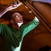 A man wearing glasses and a green long-sleeve shirt is working underneath or inside a large vehicle or aircraft, using a handheld light source to illuminate the area above him. He appears to be inspecting or performing maintenance on the underside of a large structure. The image is dramatically lit with warm amber tones contrasting with the cool green light reflecting off his shirt.