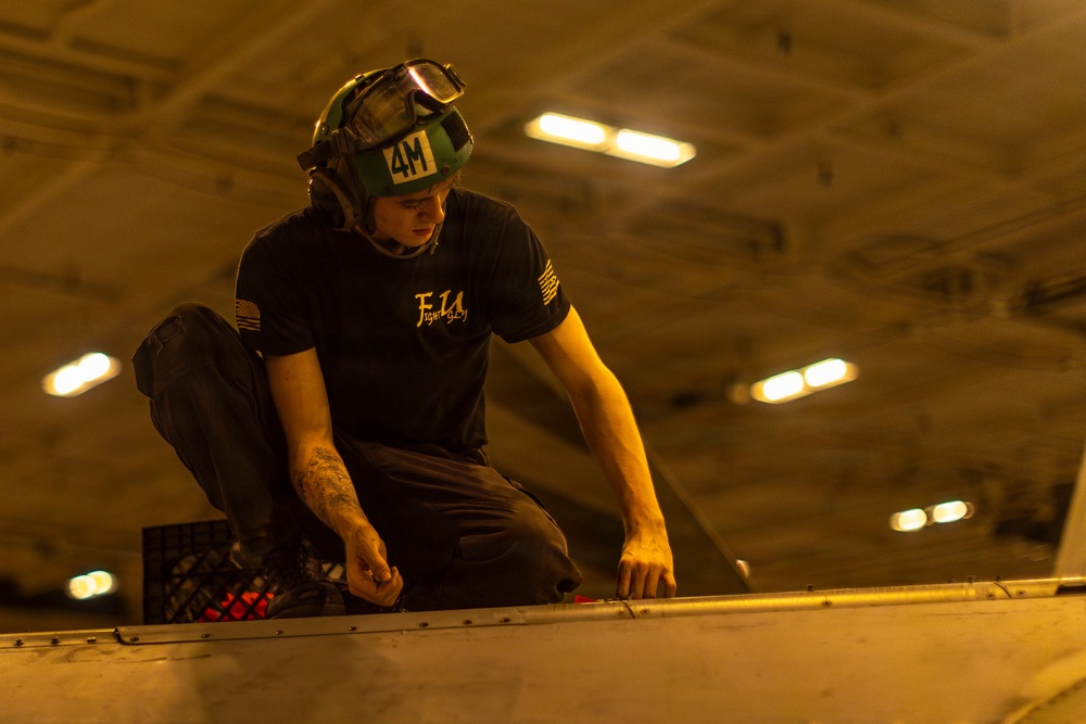 A military aviation technician or deck crew member is performing maintenance work on what appears to be an aircraft surface, likely in a carrier hangar bay. The individual is wearing a green helmet marked '4M' with goggles, a dark navy t-shirt referencing the F-14 'Tomcatters,' and has visible tattoos on their forearms. The warm amber lighting and ceiling structure are consistent with a naval aircraft carrier hangar deck environment.