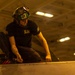 A military aviation technician or deck crew member is performing maintenance work on what appears to be an aircraft surface, likely in a carrier hangar bay. The individual is wearing a green helmet marked '4M' with goggles, a dark navy t-shirt referencing the F-14 'Tomcatters,' and has visible tattoos on their forearms. The warm amber lighting and ceiling structure are consistent with a naval aircraft carrier hangar deck environment.