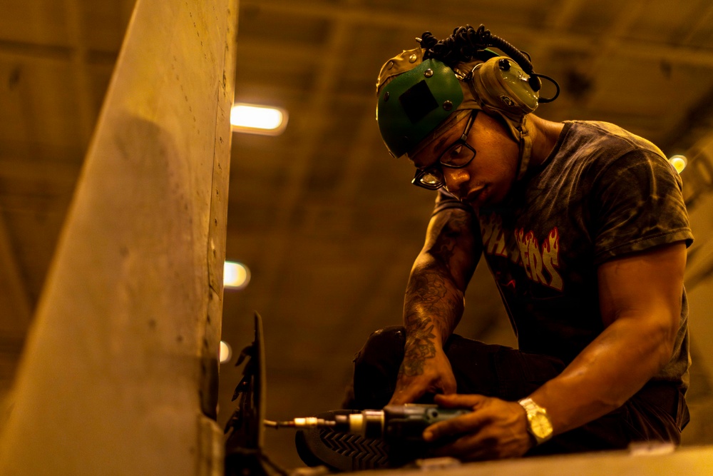 A male worker is focused on drilling or fastening something near a large flat surface, likely in an industrial or workshop environment. He is wearing personal protective equipment including ear protection and safety glasses pushed up on his forehead. The scene is lit with warm amber tones from overhead industrial lighting.