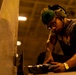 A male worker is focused on drilling or fastening something near a large flat surface, likely in an industrial or workshop environment. He is wearing personal protective equipment including ear protection and safety glasses pushed up on his forehead. The scene is lit with warm amber tones from overhead industrial lighting.