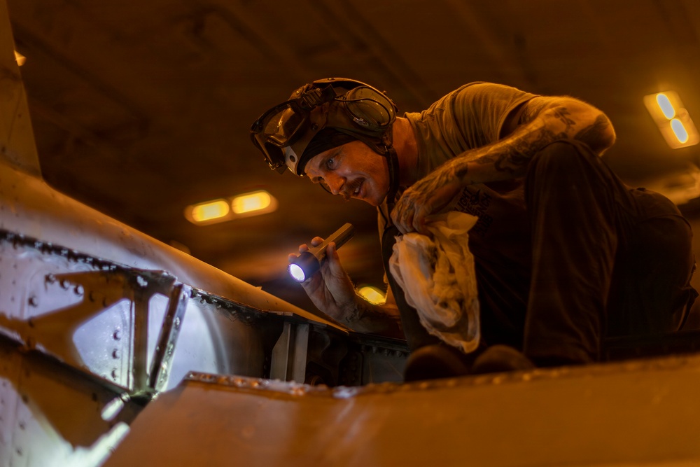 A technician or maintenance worker is inspecting a large metal structure, likely an aircraft or naval vessel component, using a handheld flashlight in a dimly lit environment. The worker is wearing protective goggles pushed up on their forehead and is leaning over riveted metal panels to examine them closely. The warm amber lighting of the surrounding space contrasts with the bright white beam of the flashlight.