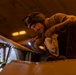 A technician or maintenance worker is inspecting a large metal structure, likely an aircraft or naval vessel component, using a handheld flashlight in a dimly lit environment. The worker is wearing protective goggles pushed up on their forehead and is leaning over riveted metal panels to examine them closely. The warm amber lighting of the surrounding space contrasts with the bright white beam of the flashlight.
