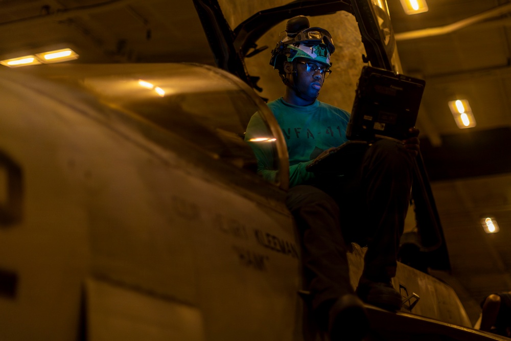 A military aviation technician or crew member is seated in or on a military aircraft cockpit in what appears to be a hangar bay, illuminated by dim ambient lighting. The individual is wearing a green shirt with 'VFA-41' visible, a helmet with night vision goggle mounts, and is reviewing a tablet or electronic device. The low-light environment and equipment suggest this is a maintenance or pre-flight inspection activity aboard a naval vessel.