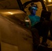 A military aviation technician or crew member is seated in or on a military aircraft cockpit in what appears to be a hangar bay, illuminated by dim ambient lighting. The individual is wearing a green shirt with 'VFA-41' visible, a helmet with night vision goggle mounts, and is reviewing a tablet or electronic device. The low-light environment and equipment suggest this is a maintenance or pre-flight inspection activity aboard a naval vessel.