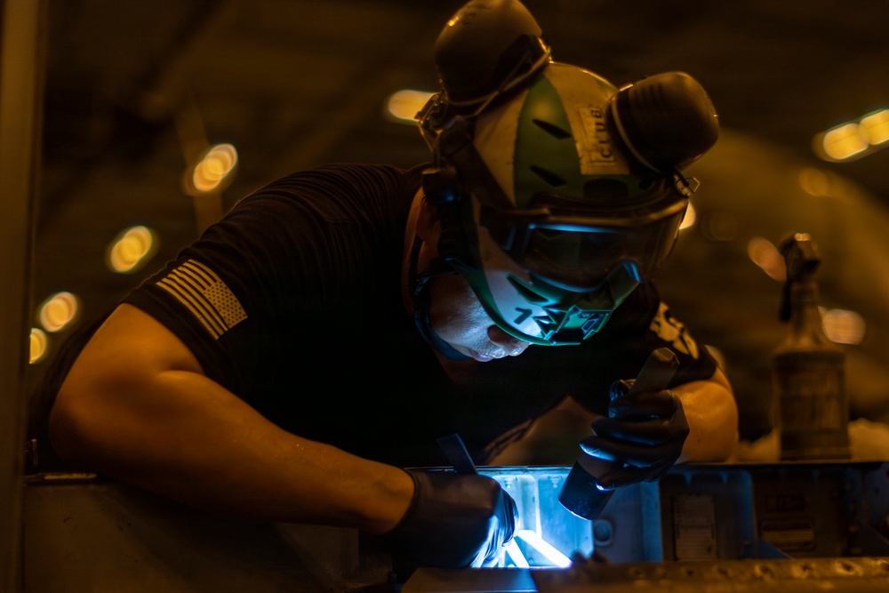 A worker wearing protective gear including a welding helmet and black gloves is performing a welding operation on a metal surface. The scene is illuminated by the bright blue-white arc light of the welding process against a dark industrial background. An American flag patch is visible on the worker's left sleeve, suggesting possible military or government affiliation.