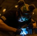 A worker wearing protective gear including a welding helmet and black gloves is performing a welding operation on a metal surface. The scene is illuminated by the bright blue-white arc light of the welding process against a dark industrial background. An American flag patch is visible on the worker's left sleeve, suggesting possible military or government affiliation.