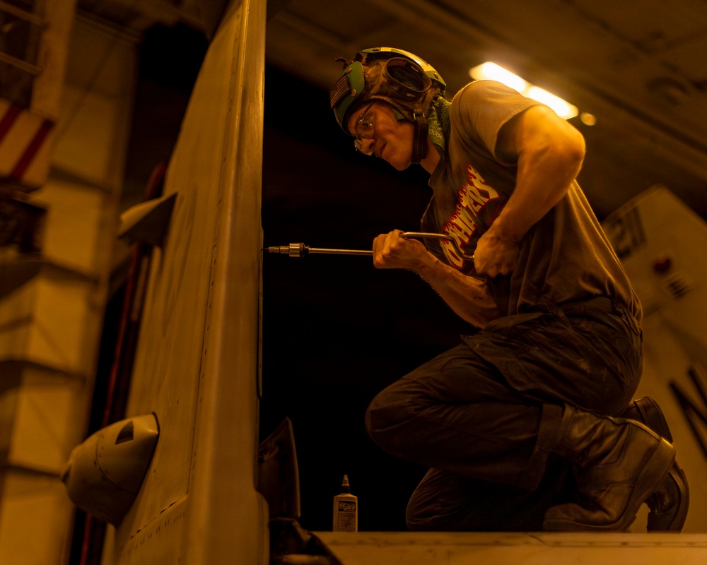 A technician or military maintainer is performing maintenance work on what appears to be a military aircraft, likely in a hangar bay. The person is kneeling on the aircraft surface and using a long-handled tool, possibly a screwdriver or torque wrench, to work on a component. The scene is illuminated by warm amber/yellow artificial lighting consistent with a ship's hangar deck environment.