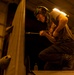 A technician or military maintainer is performing maintenance work on what appears to be a military aircraft, likely in a hangar bay. The person is kneeling on the aircraft surface and using a long-handled tool, possibly a screwdriver or torque wrench, to work on a component. The scene is illuminated by warm amber/yellow artificial lighting consistent with a ship's hangar deck environment.