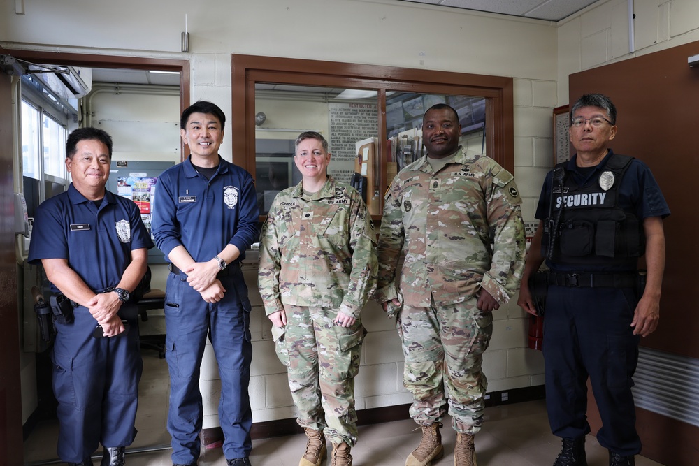 Five personnel are posing together indoors, consisting of two U.S. Army soldiers in OCP (Operational Camouflage Pattern) uniforms and three security/law enforcement personnel in navy blue uniforms. The setting appears to be a security checkpoint or guard station, likely on a military installation. The group appears to be posing for an official or commemorative photograph.
