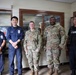 Five personnel are posing together indoors, consisting of two U.S. Army soldiers in OCP (Operational Camouflage Pattern) uniforms and three security/law enforcement personnel in navy blue uniforms. The setting appears to be a security checkpoint or guard station, likely on a military installation. The group appears to be posing for an official or commemorative photograph.