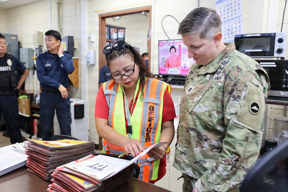 A U.S. Army soldier in OCP camouflage uniform reviews documents with a civilian officer wearing a high-visibility safety vest inside what appears to be a facility operations or security office. In the background, a security guard and another individual are visible near electrical panels and a television monitor. The scene suggests an official inspection, briefing, or coordination meeting.