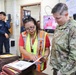 A U.S. Army soldier in OCP camouflage uniform reviews documents with a civilian officer wearing a high-visibility safety vest inside what appears to be a facility operations or security office. In the background, a security guard and another individual are visible near electrical panels and a television monitor. The scene suggests an official inspection, briefing, or coordination meeting.